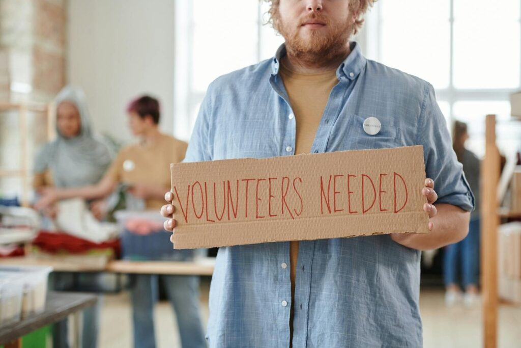 A man holds a 'Volunteers Needed' sign in a charitable organization setting, highlighting the need for help.