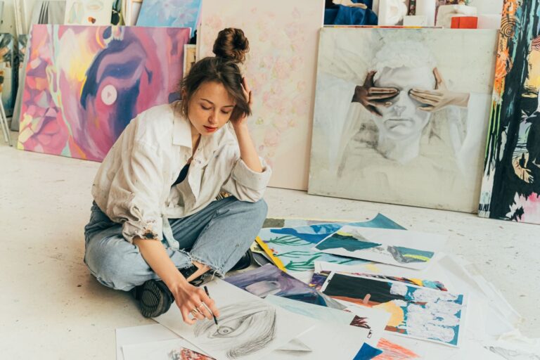 A young woman artist sketching on the floor surrounded by paintings in a creative studio.