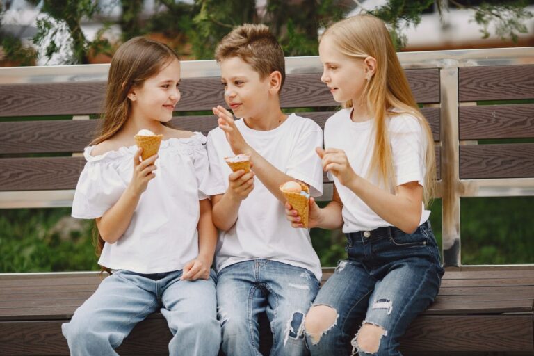 Three happy children sitting on a bench enjoying ice cream cones together outdoors.