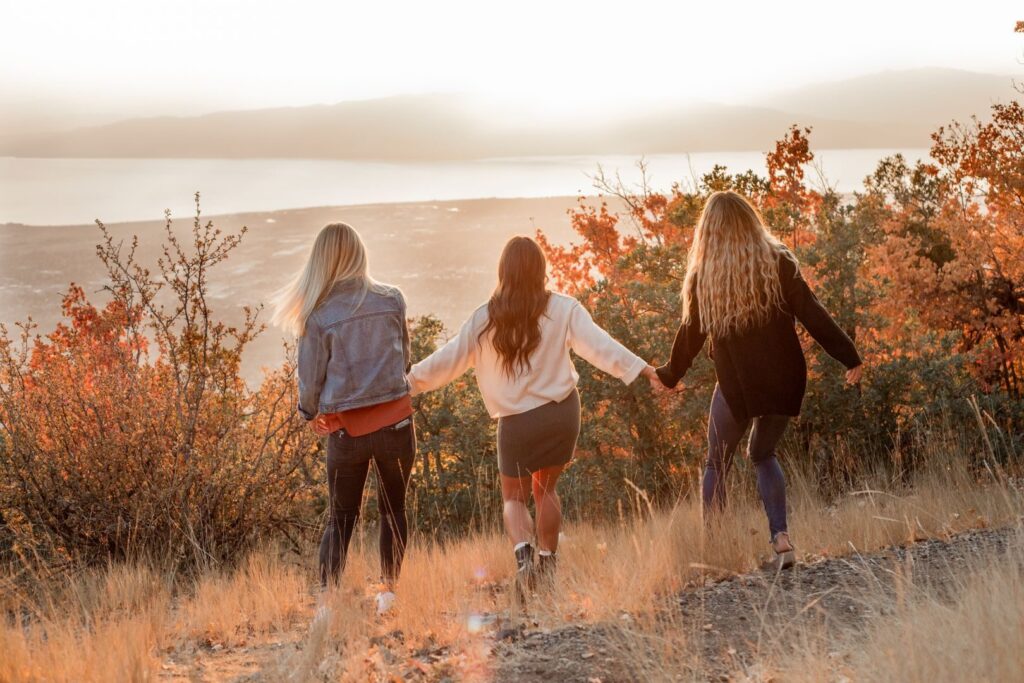 Three women hike together in a scenic autumn landscape, holding hands and enjoying nature.